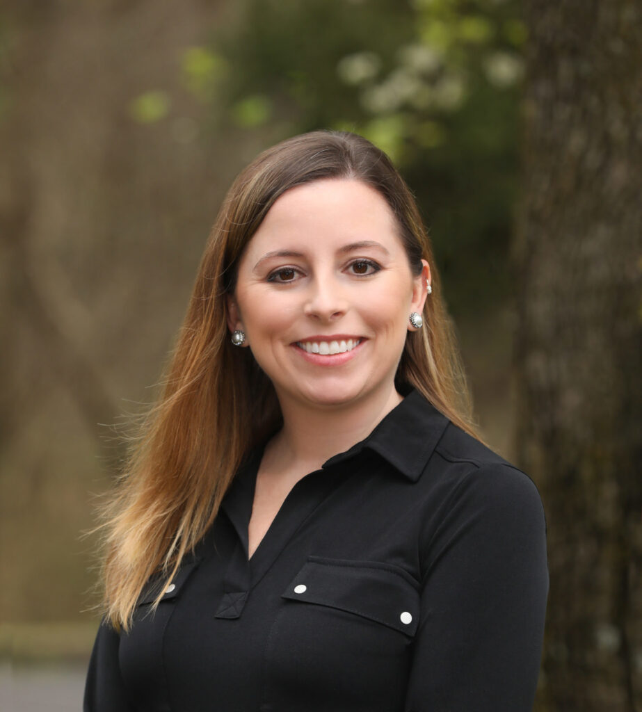 A woman with straight, light brown hair wearing a black button-up shirt stands outdoors, smiling at the camera with a blurred background of trees.