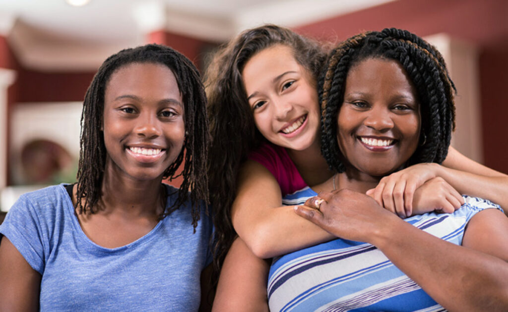 Three women smile at the camera indoors. One woman hugs another from behind while the third sits beside them. All appear happy and relaxed in a home setting.