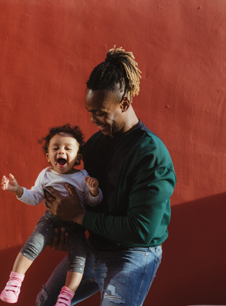 An adult holds a smiling toddler in front of a red wall. The toddler appears happy, wearing a white shirt and gray pants; the adult is in a green sweater and ripped jeans.