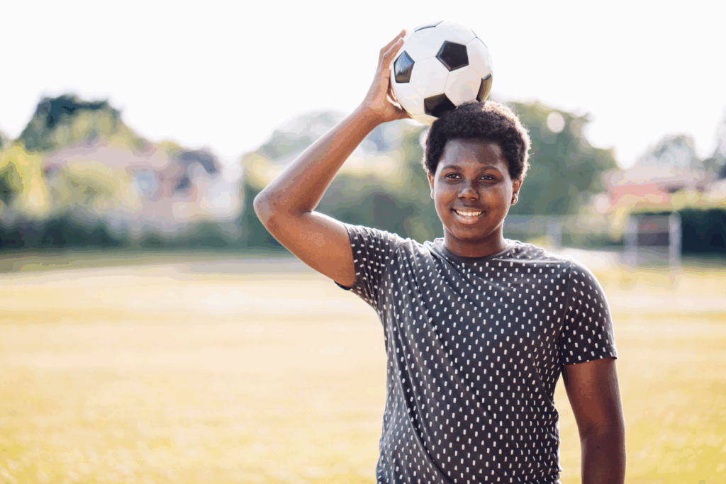A young person standing outdoors on a soccer field holds a soccer ball on their head and smiles at the camera.