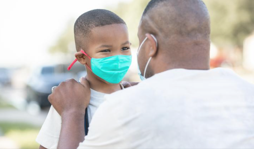 An adult facing a young boy outside, both wearing protective face masks. The adult holds the boy by the shoulders as they make eye contact.