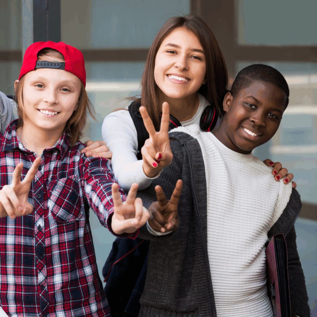 Three smiling kids stand close together, making peace signs with their fingers. One wears a red cap backward, another has long hair, and the third holds a folder.