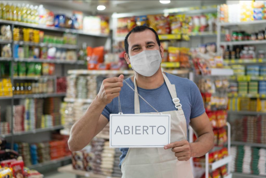 A man wearing a face mask and apron stands in a grocery store aisle holding a sign that says "Abierto," indicating the store is open.