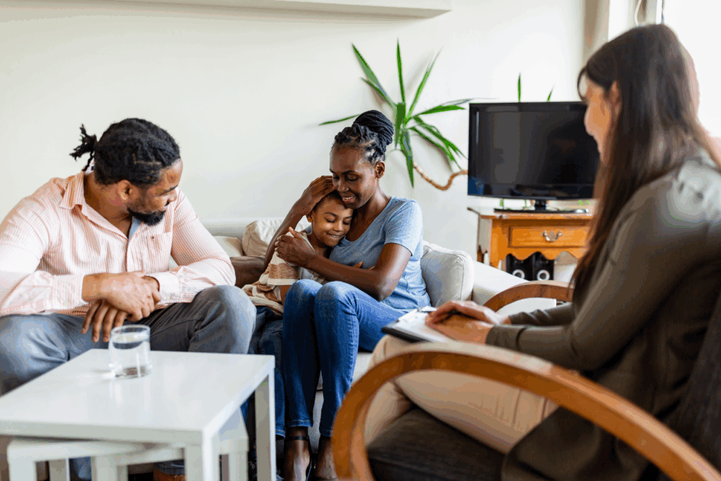 A family sits together on a couch, with a woman hugging a child, while another woman takes notes nearby, suggesting a counseling or therapy session.
