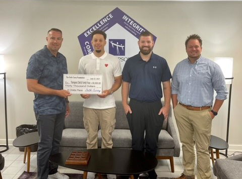 Four men stand indoors, two holding a large check for $30,000, in front of a wall sign displaying the words "EXCELLENCE," "INTEGRITY," and "COMMUNITY.
