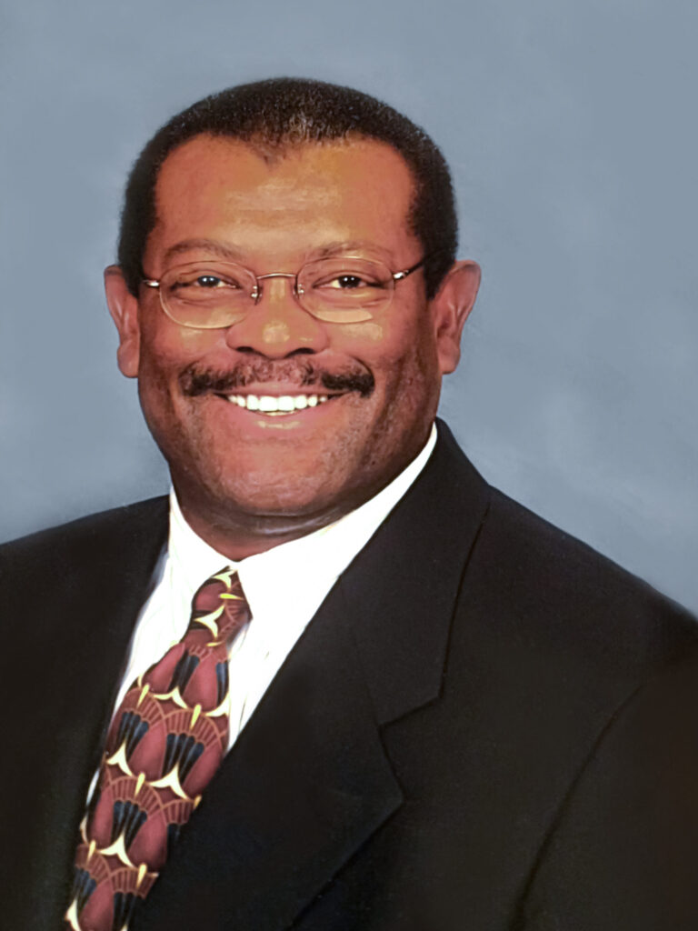 A man wearing glasses, a dark suit, a white shirt, and a patterned tie, smiling in front of a plain blue background.