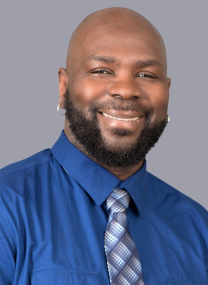A man with a beard and shaved head, wearing a blue dress shirt and patterned tie, smiles in front of a plain gray background.
