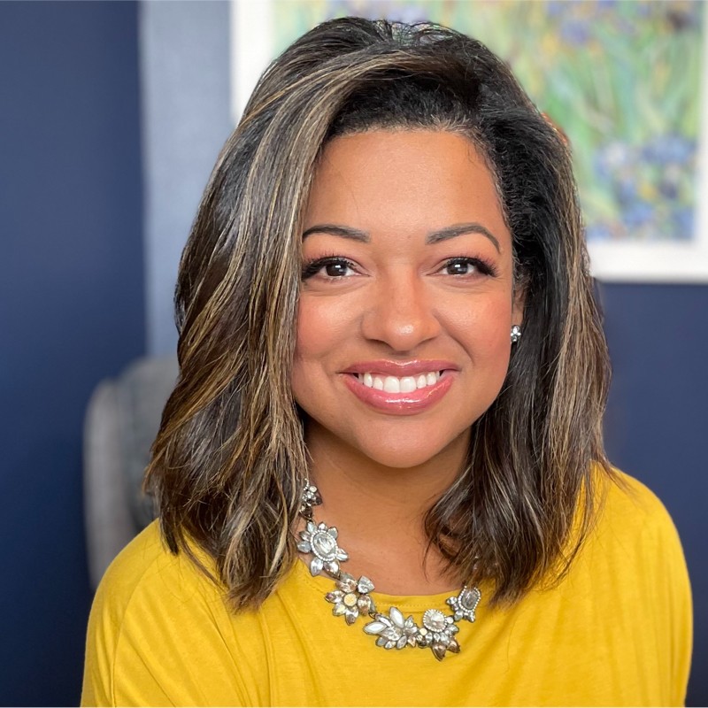 Woman with shoulder-length brown hair wearing a yellow top and a floral necklace, smiling in front of a navy blue wall with a framed artwork.