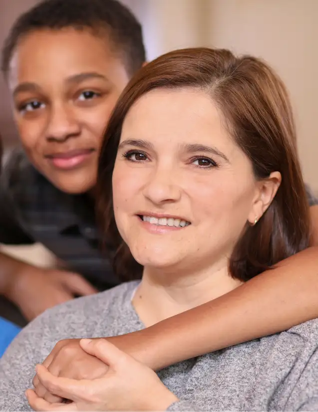 A woman sits and smiles at the camera while a boy stands behind her with his arm gently resting on her shoulder.