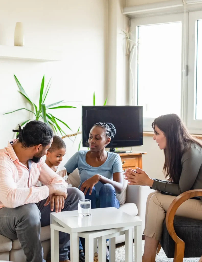 A family of three sits on a couch talking with a woman who appears to be a counselor in a bright living room.