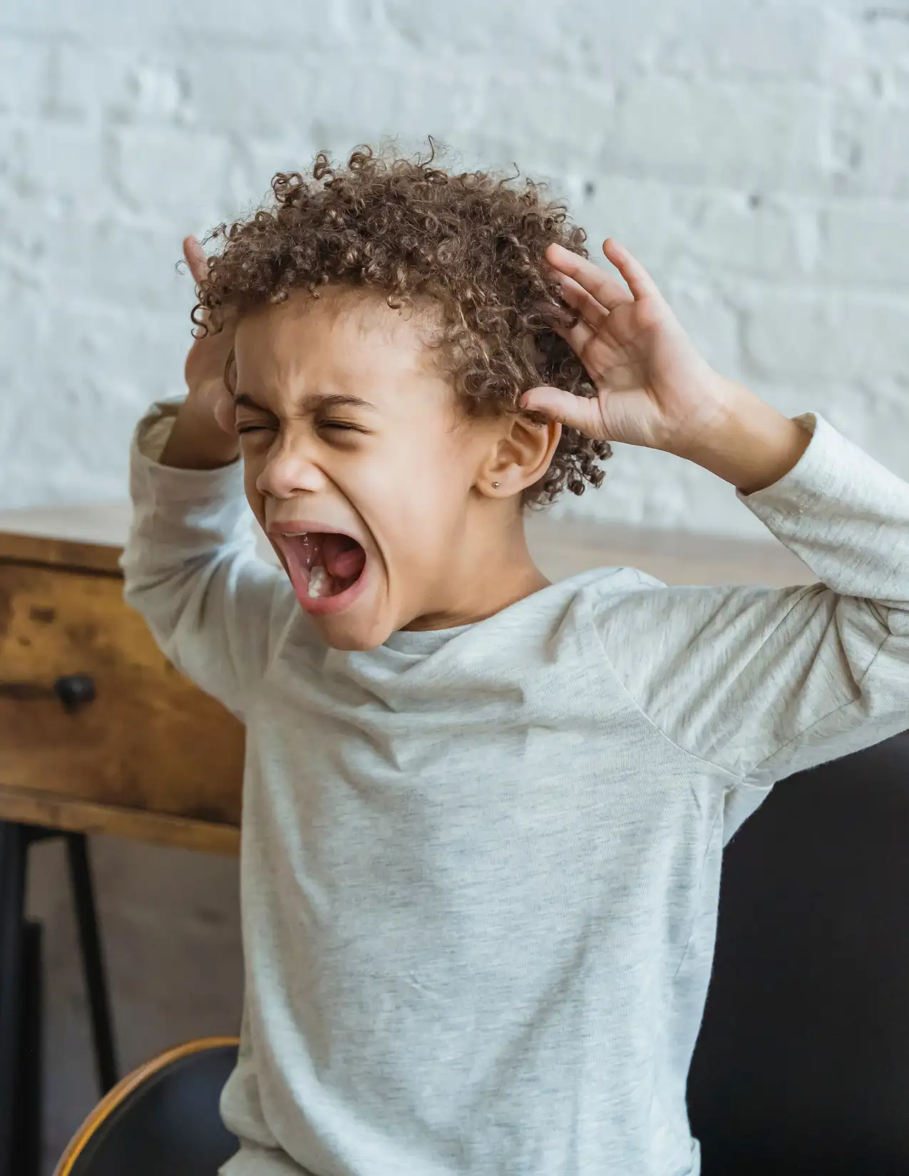 A young child with curly hair, wearing a light gray shirt, sits on a chair and screams with eyes closed and hands raised near their head.