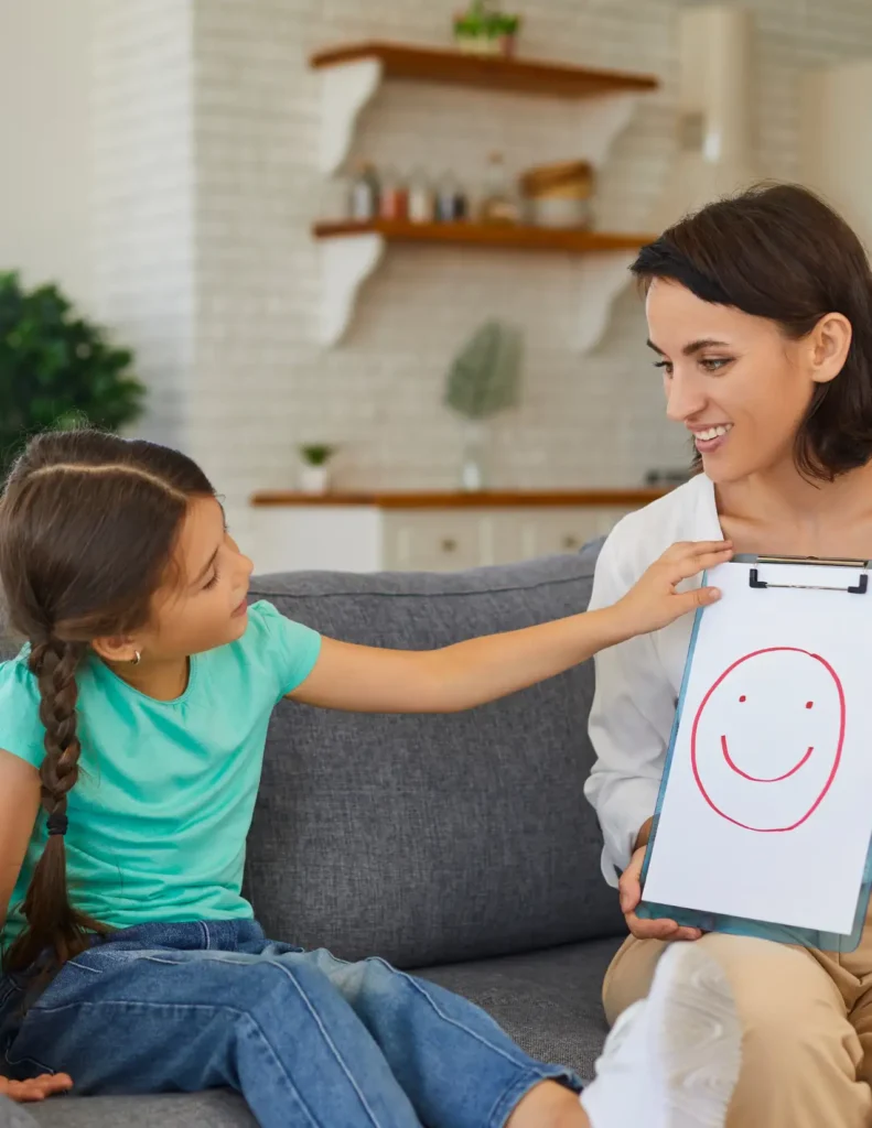 A woman holds a clipboard with a drawing of a smiling face while a young girl with braided hair points at it. They are sitting on a couch in a living room.