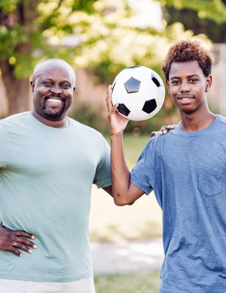 Two people stand outdoors; one holds a soccer ball and both are smiling at the camera. Trees and sunlight are visible in the background.