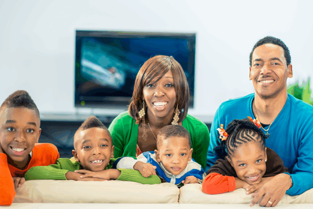 A family of five, including two adults and three children, sits together on a couch and smiles at the camera with a television in the background.