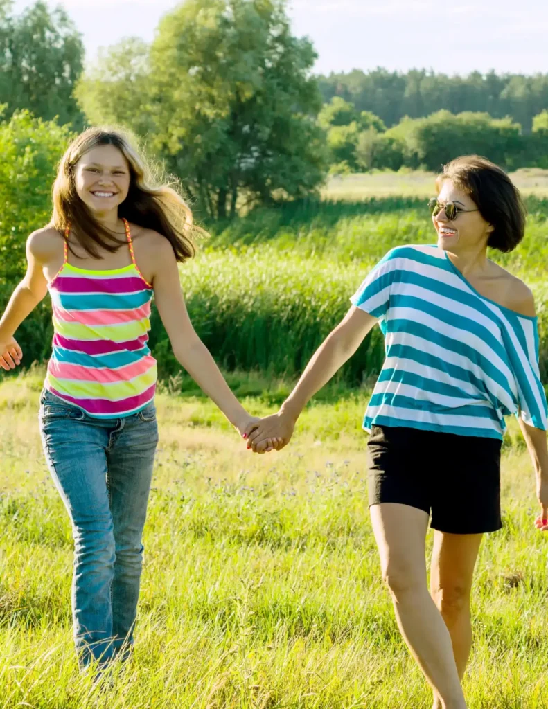 Two women are holding hands and walking in a grassy field on a sunny day, both smiling and wearing colorful striped tops. Trees and greenery are visible in the background.
