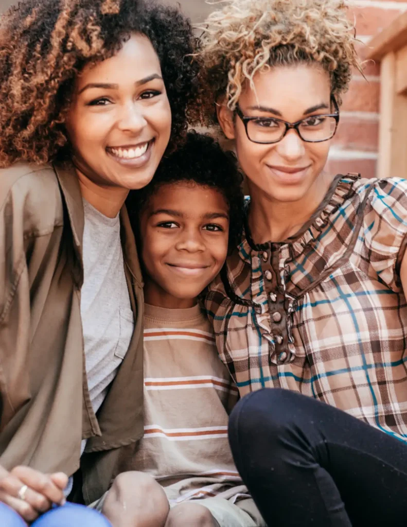 Two women and a boy sit closely together, smiling at the camera. One woman wears glasses and a plaid shirt; the other wears a jacket. The boy is in a striped t-shirt.