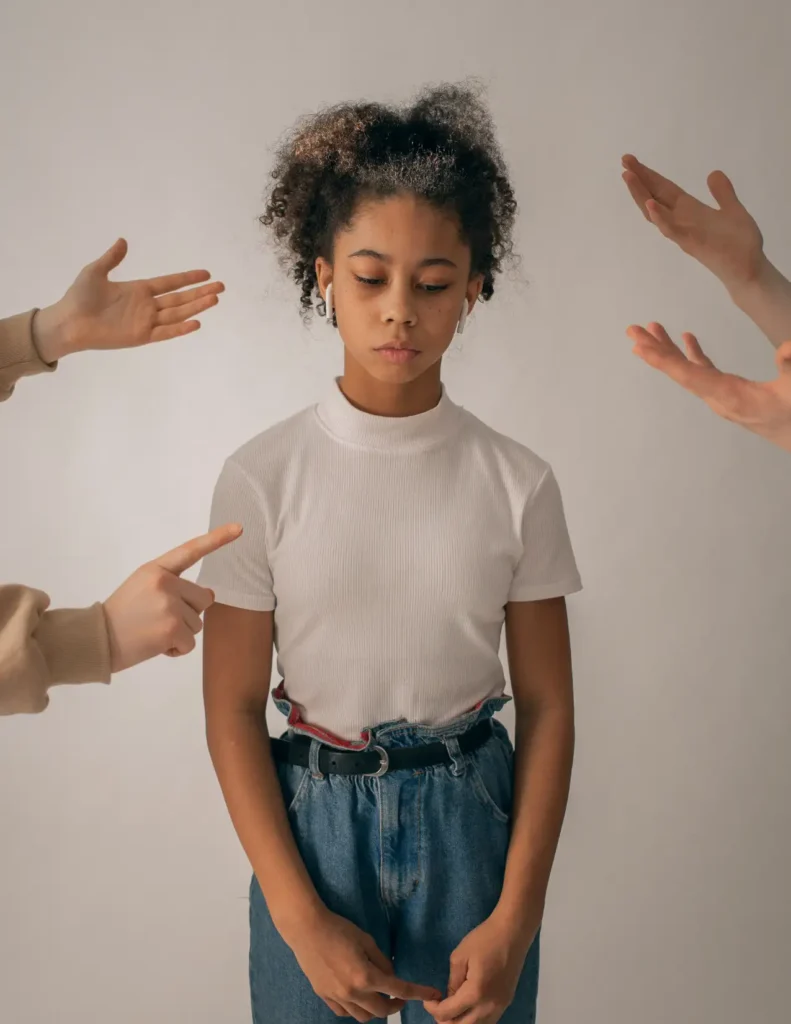 A young girl stands with her head down, surrounded by adult hands pointing and gesturing towards her against a plain background.
