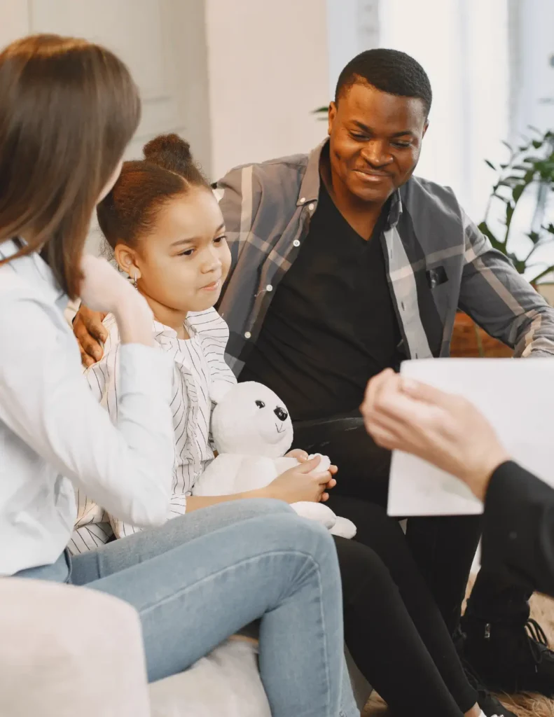 A young girl holding a stuffed animal sits between two adults on a couch, listening to another person who is holding a clipboard.
