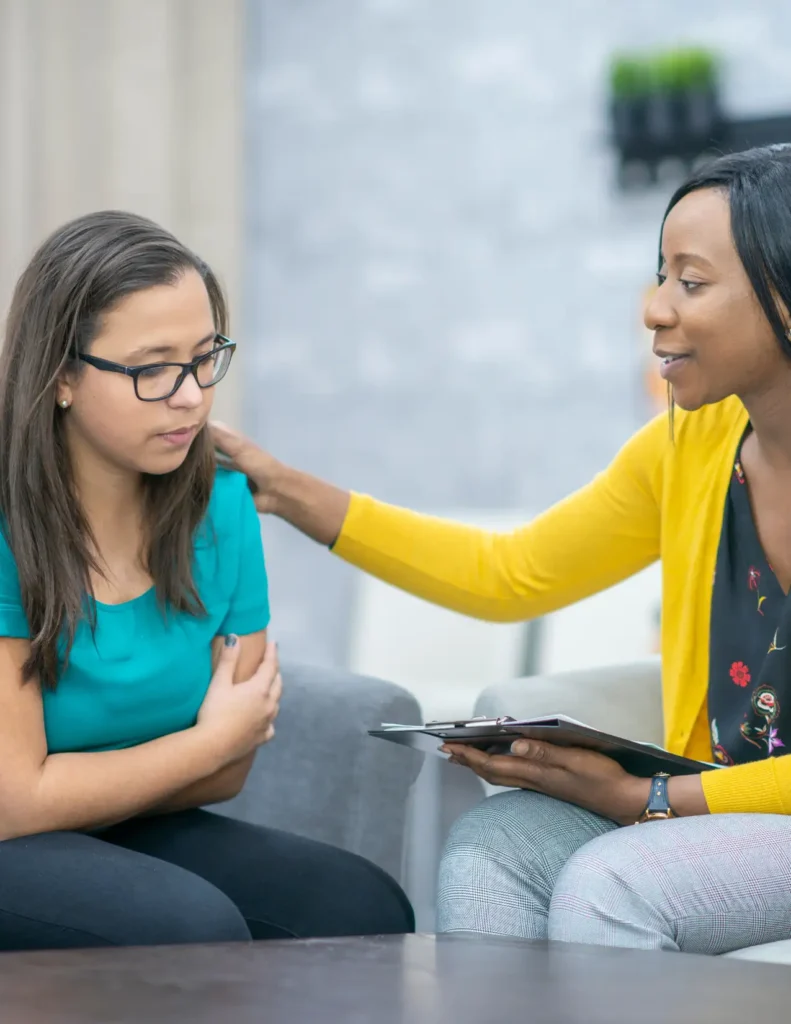 A woman in a yellow sweater comforts a young woman in glasses who appears upset, while holding a clipboard during a counseling session.