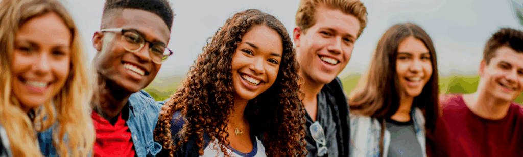 A group of six teenagers stands closely together outdoors, smiling and looking at the camera.