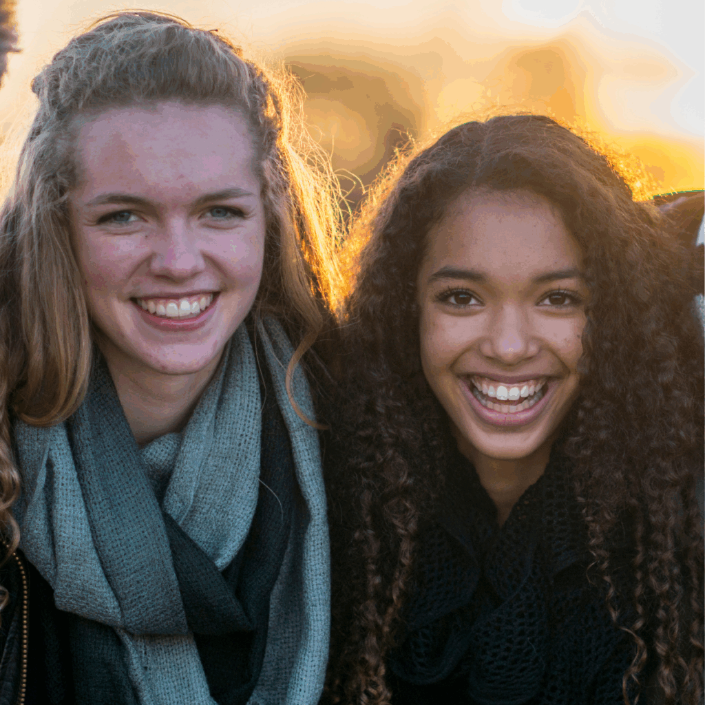 Two young women wearing scarves stand close together outdoors, both smiling at the camera with sunlight in the background.