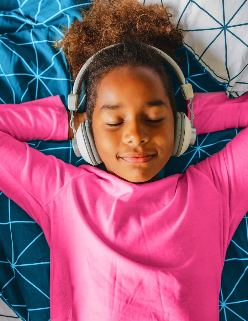 A child wearing a bright pink shirt and headphones is lying on a blue geometric-patterned bedspread with eyes closed and arms behind their head.