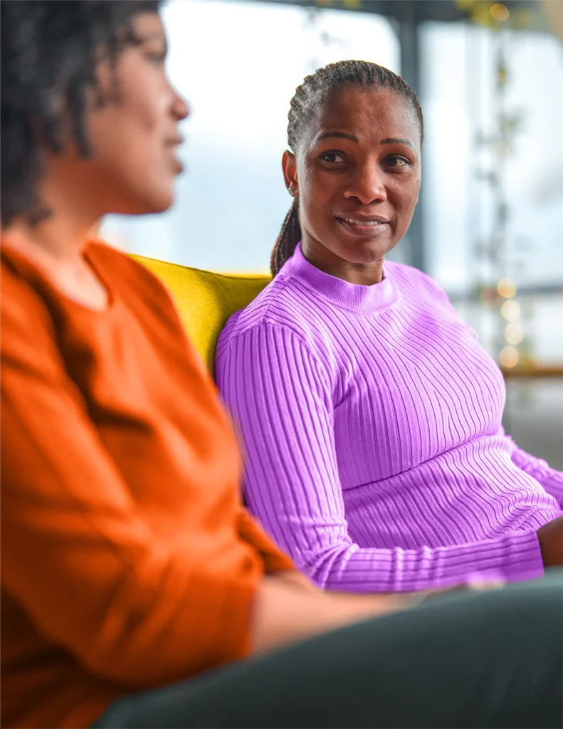 Two women are sitting and talking indoors; one wears an orange top, while the other, facing her, wears a light purple ribbed sweater and listens attentively.