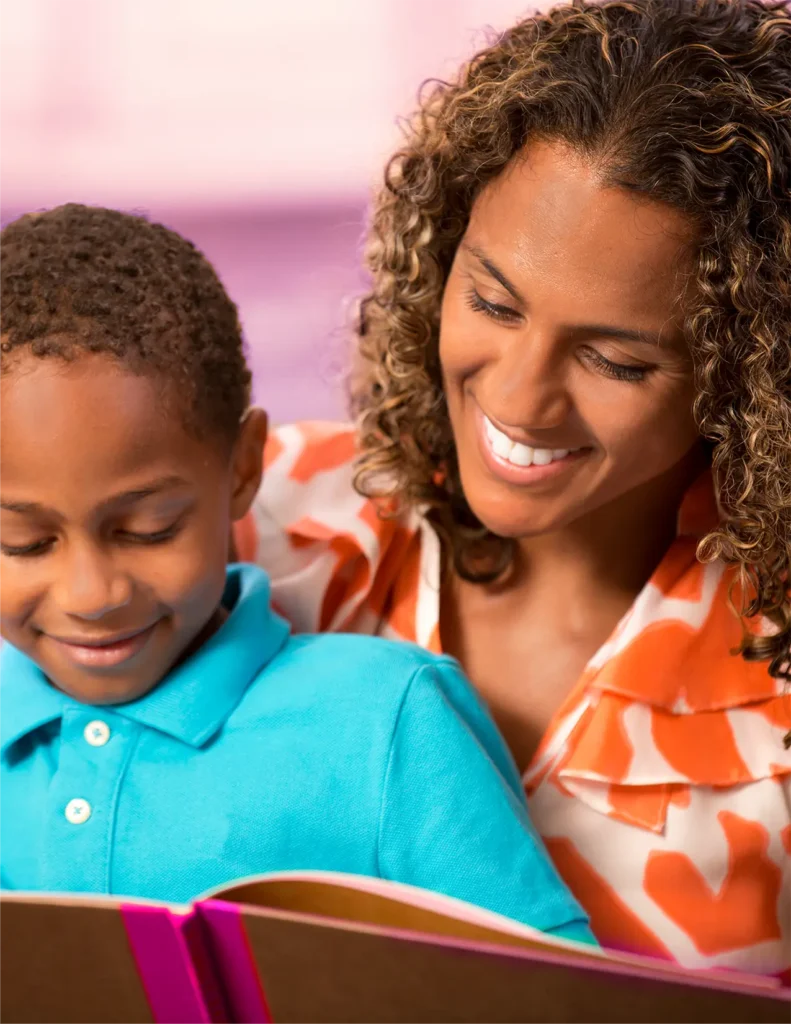 An adult and a child sit close together, smiling as they read a book with a pink spine. The child wears a blue shirt; the adult wears an orange and white top.