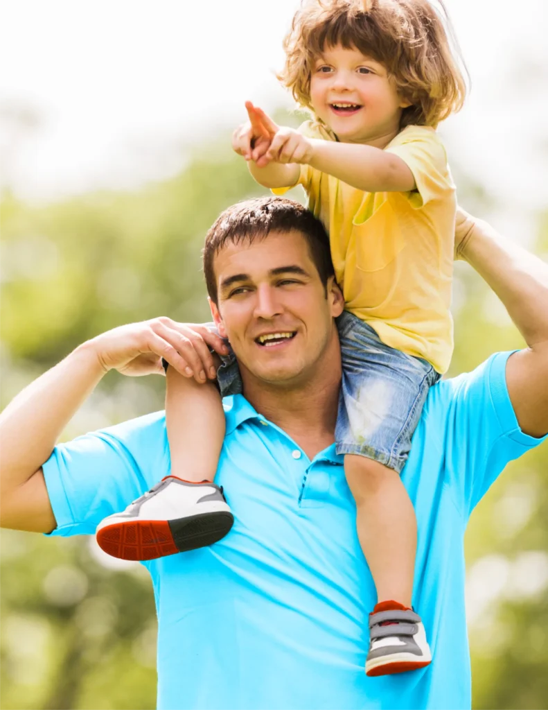 A man in a blue shirt carries a young child on his shoulders outdoors; the child is smiling and pointing ahead.