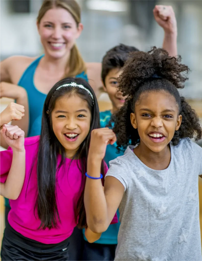 A group of children and a woman pose together indoors, smiling and flexing their arms in a gesture of strength.