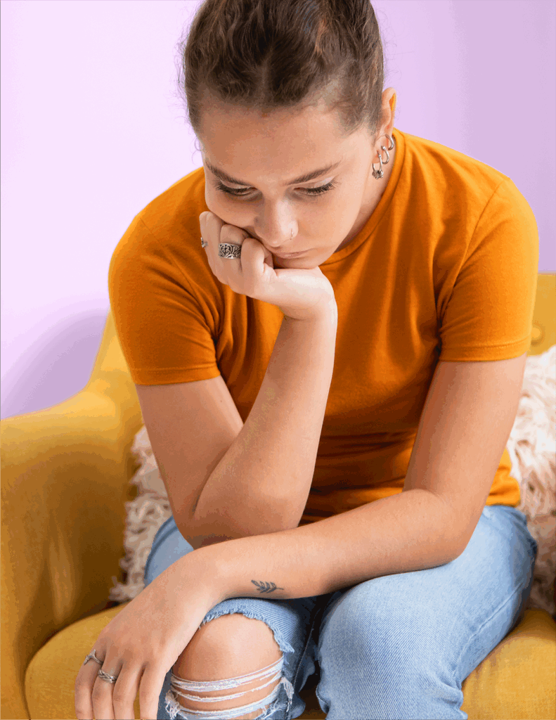 A young person in an orange shirt and ripped jeans sits on a yellow chair, looking down with a thoughtful expression and resting their chin on their hand.