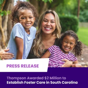 A woman sits outdoors smiling with two young girls, one on each side of her. Text overlay announces funding for foster care services in South Carolina.