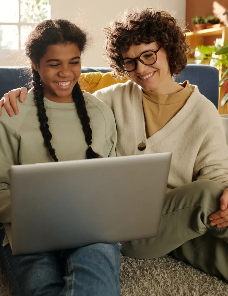 Two people sit closely together on a couch, smiling as they look at a laptop screen in a cozy, sunlit living room.