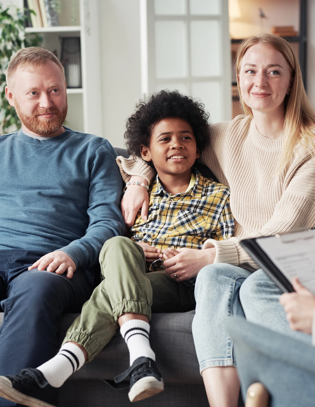 A man, woman, and child sit together on a couch, facing someone holding a clipboard, suggesting a family meeting or counselling session.