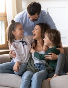 A man, woman, and two children sitting together on a couch, smiling and laughing while embracing each other in a bright living room.