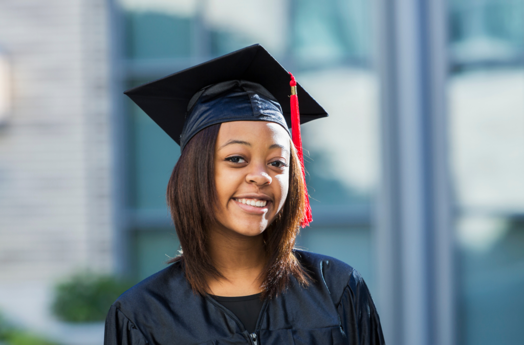 Young woman wearing a black graduation cap and gown, standing outdoors and smiling at the camera.