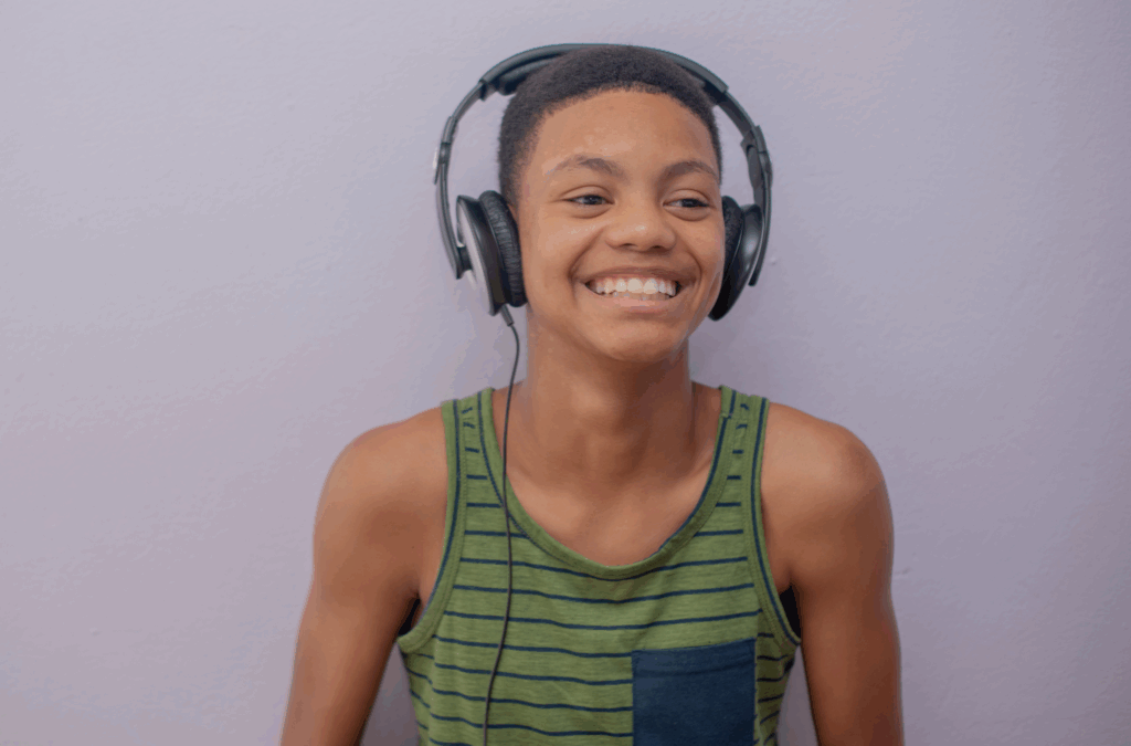 Smiling young person in a green striped tank top wearing large headphones, standing against a plain light purple wall.