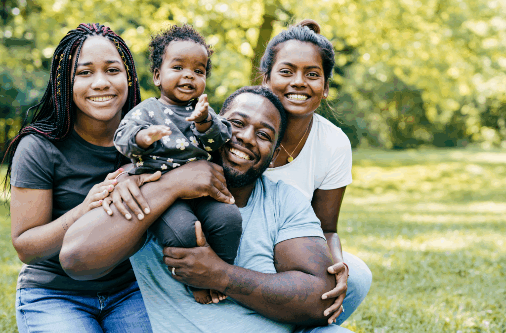 A smiling family of four poses outdoors in a grassy park, with trees in the background. The father holds a baby, while two women stand close beside them.