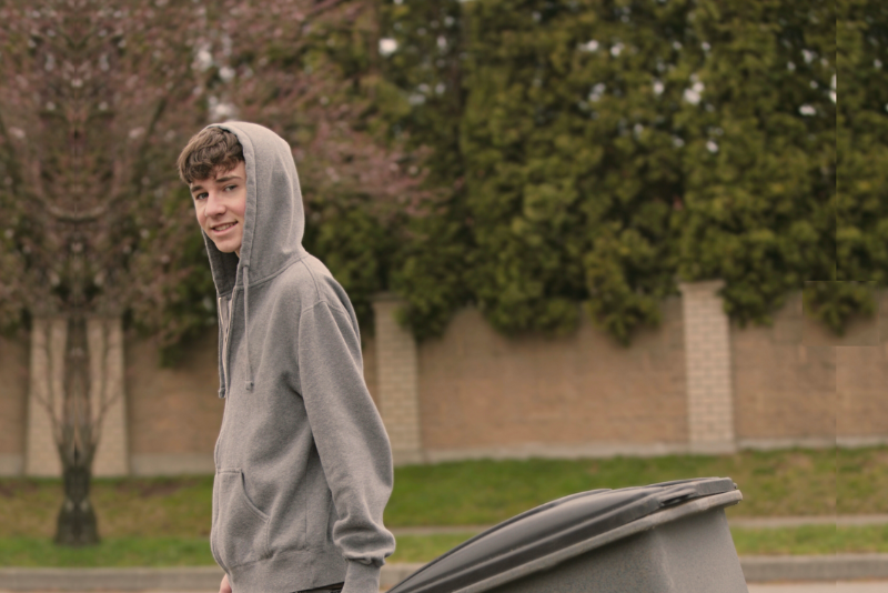 A young person in a gray hoodie pulls a large trash bin outdoors, with a brick wall and trees in the background.
