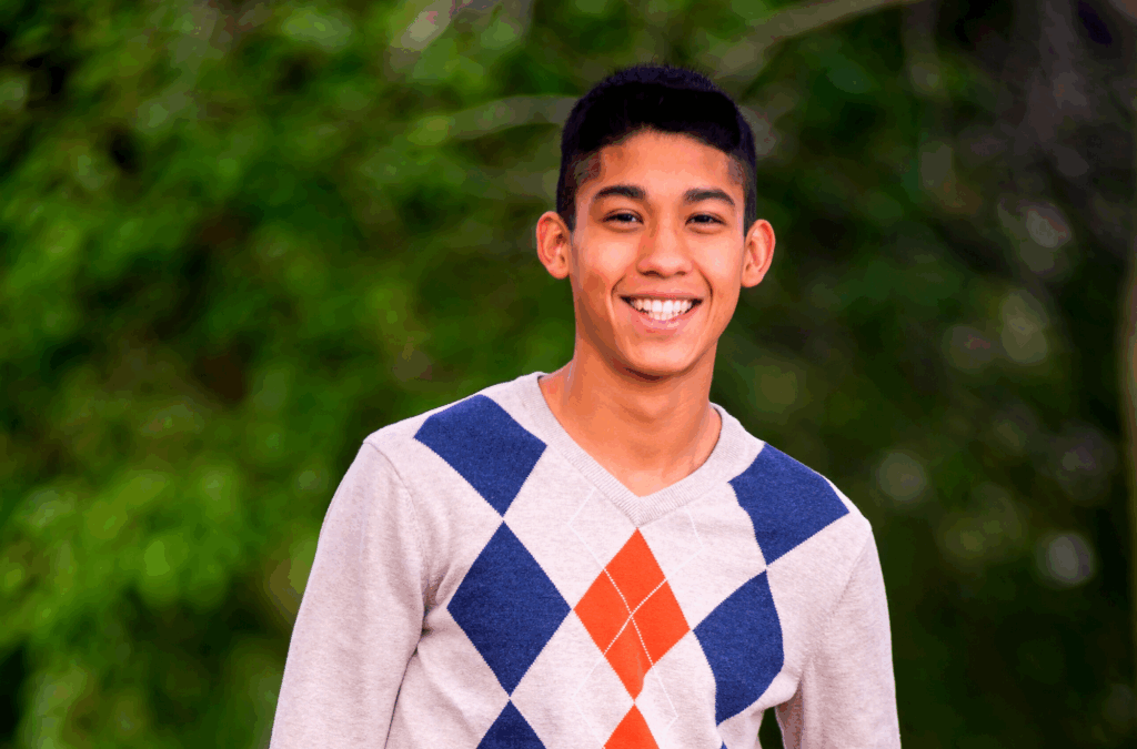 A young man with short dark hair smiles outdoors, wearing a light sweater with a blue and orange argyle pattern. Green foliage is blurred in the background.