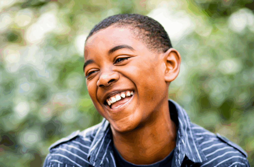 A boy with short hair and a striped shirt smiles and laughs outdoors, with a blurred green background.
