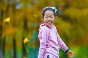 A young girl in a pink sweater smiles outdoors with autumn-colored trees and falling leaves in the background.