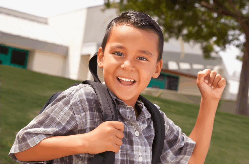 Smiling boy wearing a plaid shirt and backpack raises his fist in excitement, standing outside near a school building on a sunny day.