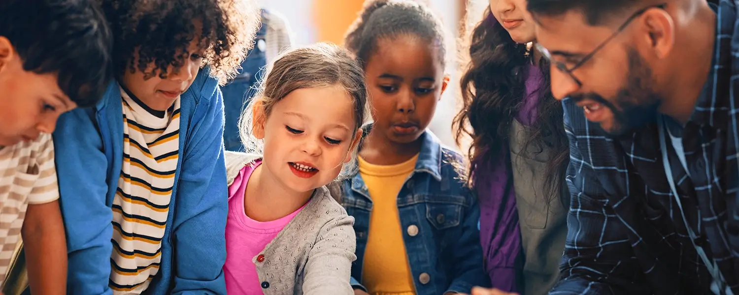 A group of young children and an adult sit closely together, engaging in an activity and looking down at something on a table.