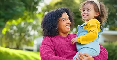 An adult and a young child are outdoors, smiling and looking at each other. The adult is holding the child, and trees are visible in the background.