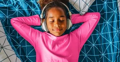 A child in a pink shirt lies on a blue patterned bed with headphones on, eyes closed and hands behind their head, appearing relaxed.