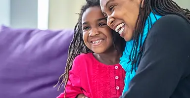An adult and a child with long hair sit close together on a couch, both smiling and appearing happy.