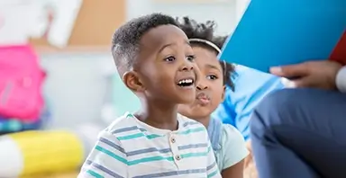 Two young children sit and listen attentively to an adult reading from a large blue book.