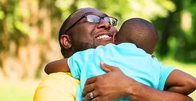A man wearing glasses smiles while hugging a young child outdoors on a sunny day.