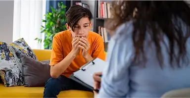 A teenage boy sits on a yellow couch, looking concerned, while a person with a clipboard faces him, suggesting a counseling or therapy session.